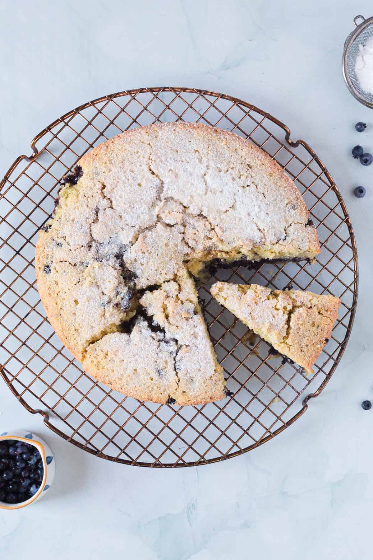 A round Blueberry Polenta Cake with a sugary top cools on a wire rack. One slice is cut and pulled out slightly. A small bowl of blueberries and a bowl of sugar sit nearby on the light surface.