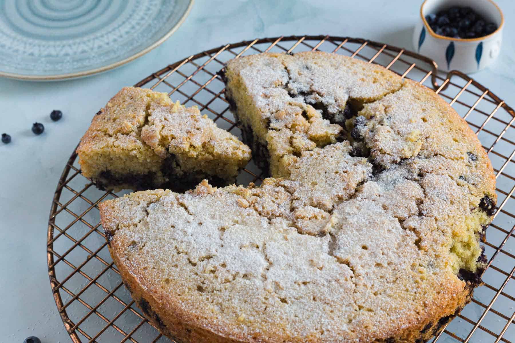 A round, sugar-dusted Blueberry Polenta Cake sits on a wire cooling rack with one slice cut and slightly separated. A small bowl of blueberries and an empty plate are nearby on a light surface.