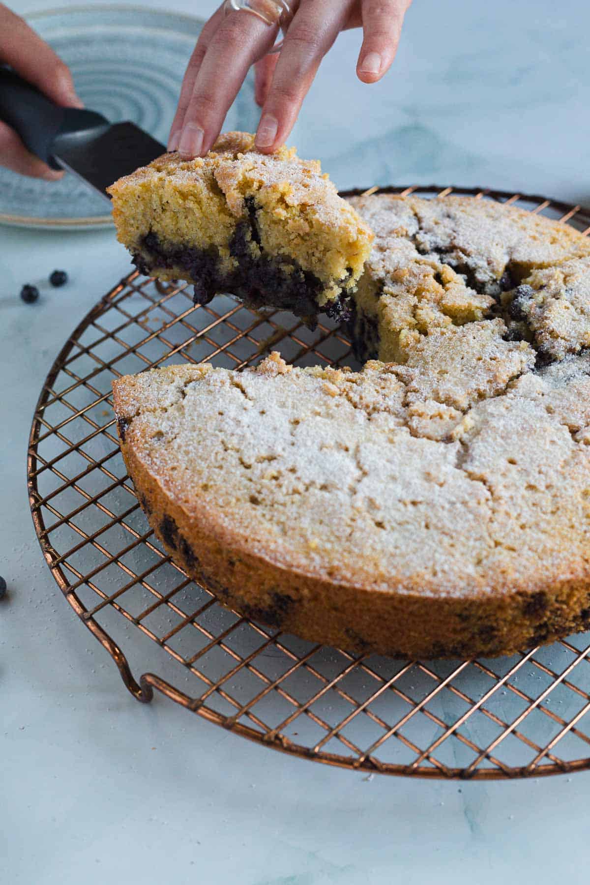 A hand lifts a slice of Blueberry Polenta Cake from a round cake resting on a wire cooling rack. The cake is dusted with sugar and shows visible berries inside. A cake server is partially visible under the lifted slice.
