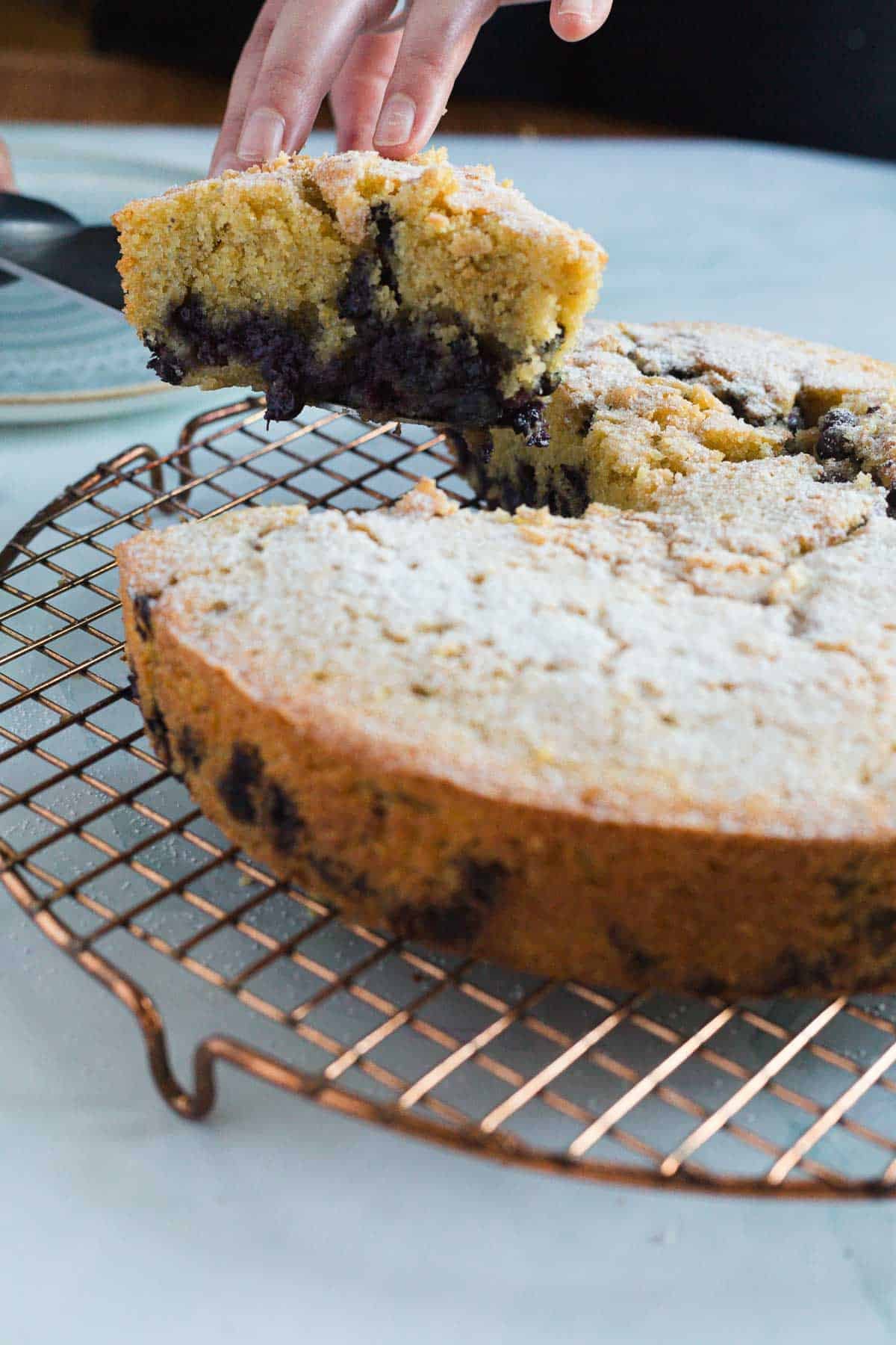 A hand lifts a slice of Blueberry Polenta Cake from a round cake on a wire cooling rack. The cake is dusted with powdered sugar and has visible blueberries inside.