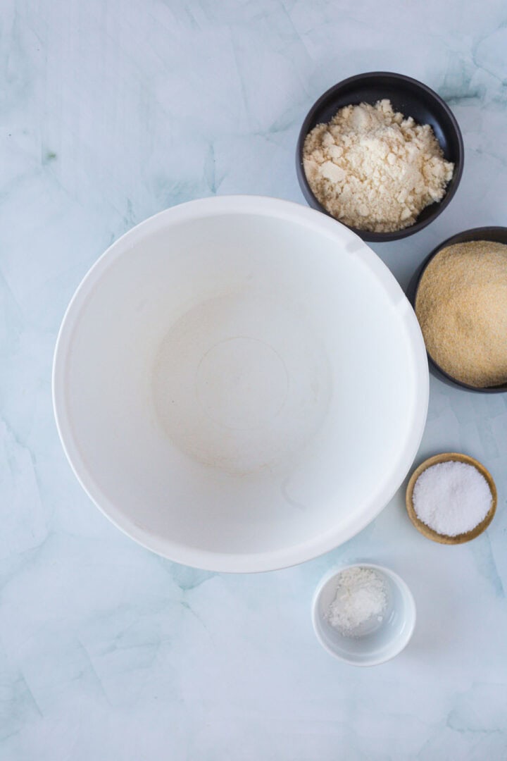 A white mixing bowl is placed on a marble surface, surrounded by smaller bowls holding flour, cornmeal, salt, and baking powder—the essential ingredients for a delicious Blueberry Polenta Cake.