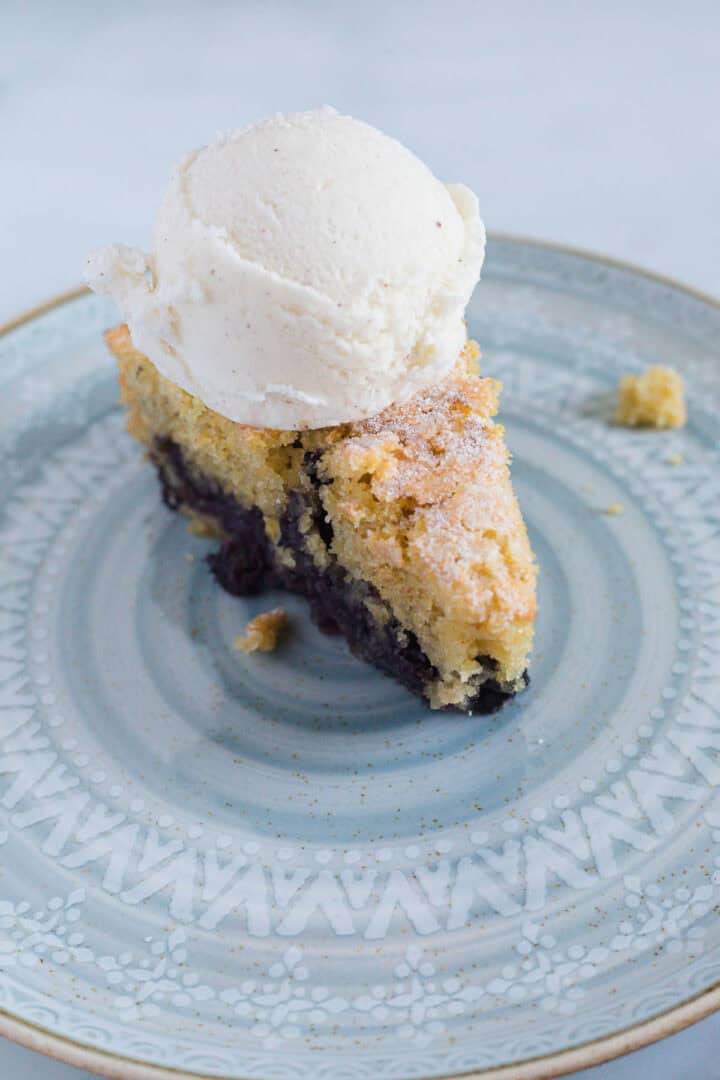 A slice of fruit cobbler with a golden crust sits on a decorative light blue plate, reminiscent of a classic Blueberry Polenta Cake, and is topped with a scoop of vanilla ice cream.