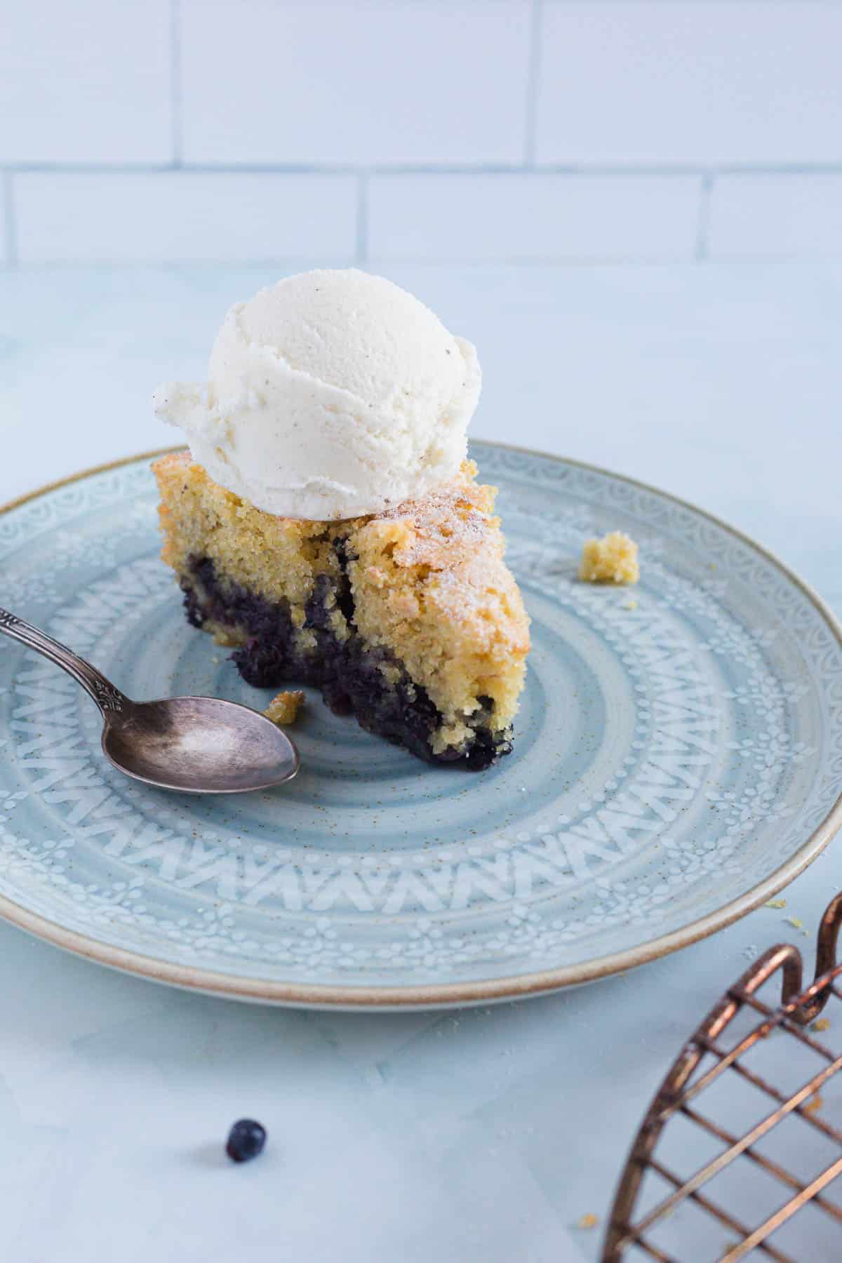 A slice of Blueberry Polenta Cake topped with a scoop of vanilla ice cream sits on a patterned plate with a spoon beside it. The surface underneath is light-colored, and a cooling rack is partially visible.