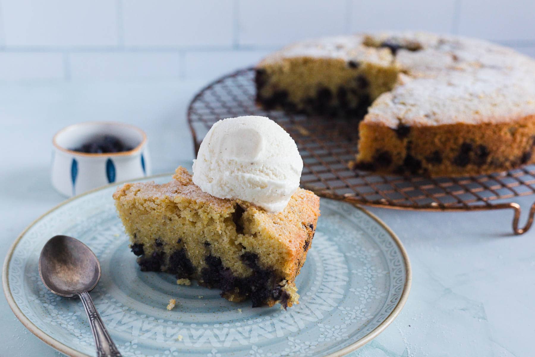 A slice of Blueberry Polenta Cake with visible chocolate chips is topped with a scoop of vanilla ice cream on a patterned plate next to a spoon. The rest of the cake sits on a cooling rack in the background.