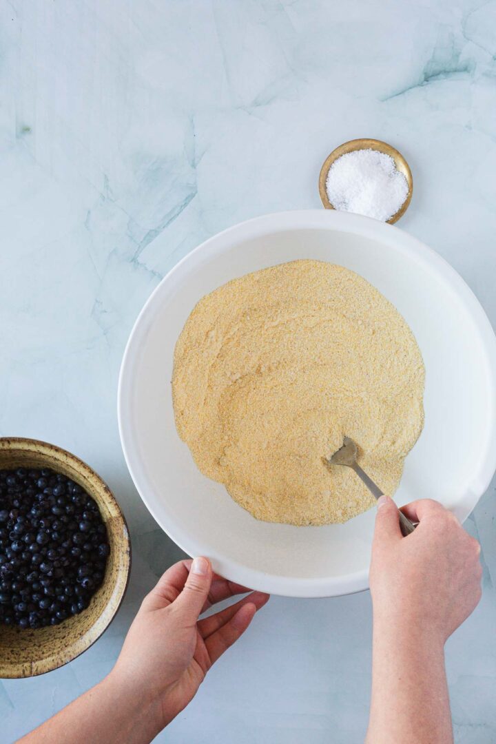 A person stirs cornmeal for a Blueberry Polenta Cake using a fork. Nearby, a bowl of blueberries and a small dish of salt rest on a light marble surface.