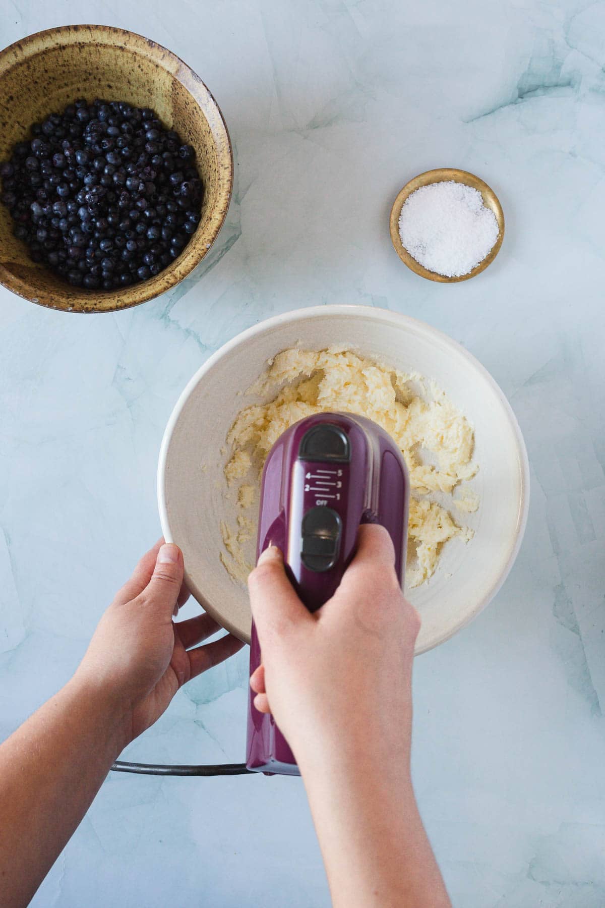 A person holds a hand mixer, creaming ingredients in a white bowl for Blueberry Polenta Cake. Beside the bowl are a small dish of salt and a larger bowl filled with blueberries on a light-colored, marble-patterned surface.