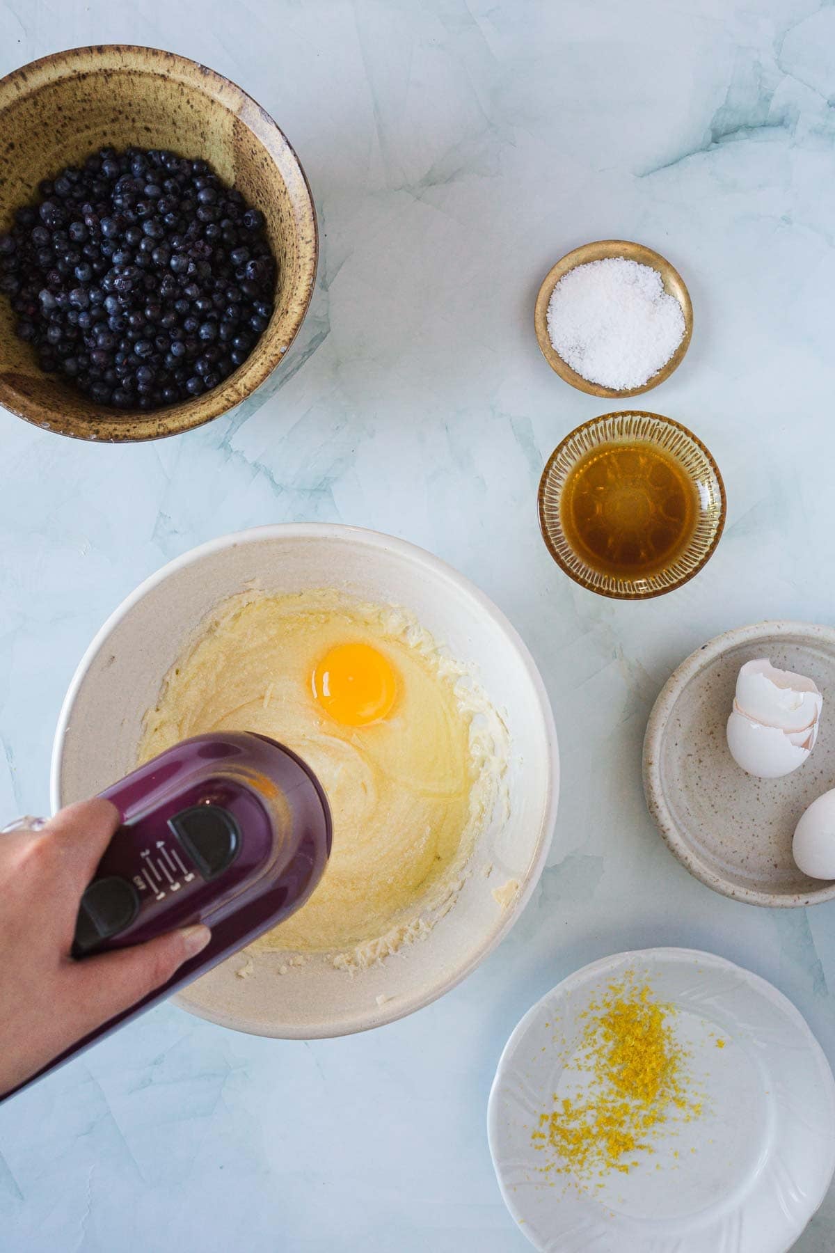 A hand uses a hand mixer to blend an egg into batter for Blueberry Polenta Cake. Around the bowl are blueberries, salt, vanilla, eggshells, lemon zest on a plate, and a bowl of sugar, all on a light countertop.