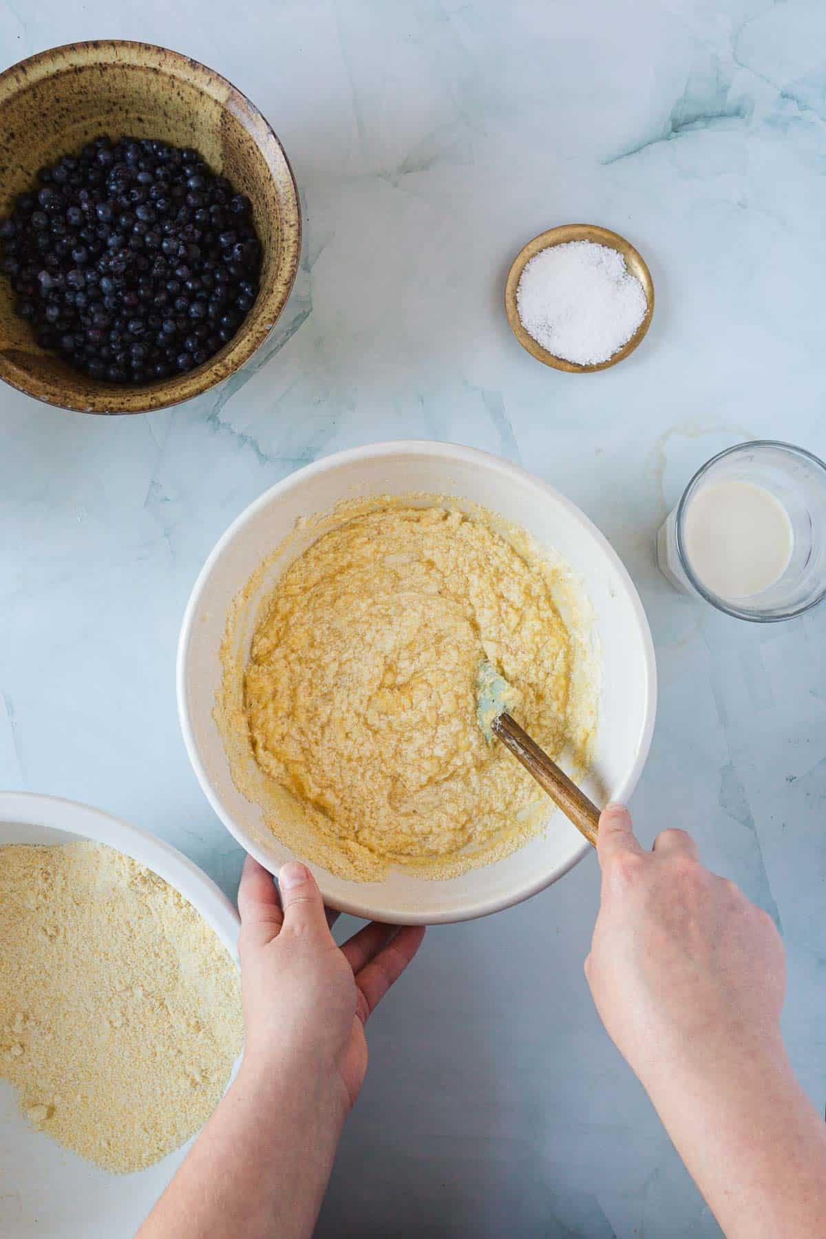 A person stirs a thick batter for Blueberry Polenta Cake in a white bowl using a spatula. Nearby, there is a bowl of blueberries, a bowl of flour, a small dish of salt, and a glass of milk on a light-colored surface.