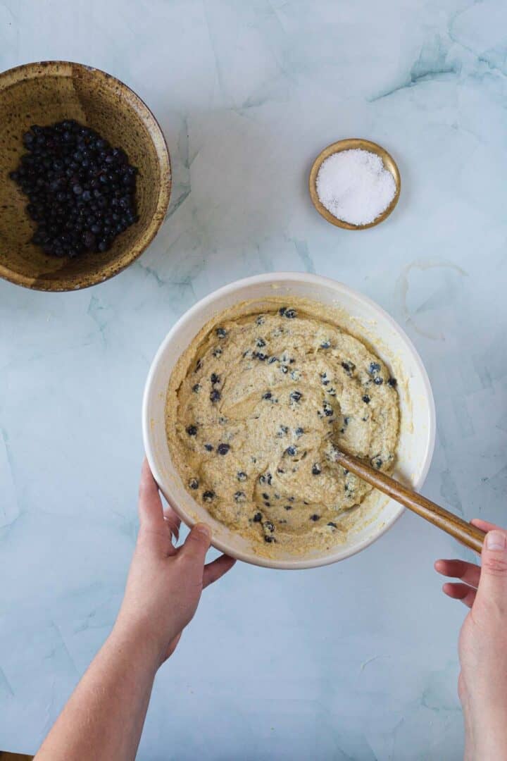 A person stirs a bowl of batter, preparing a Blueberry Polenta Cake with juicy blueberries. Nearby, a bowl of fresh blueberries and coarse sugar rest on a light blue marble surface.