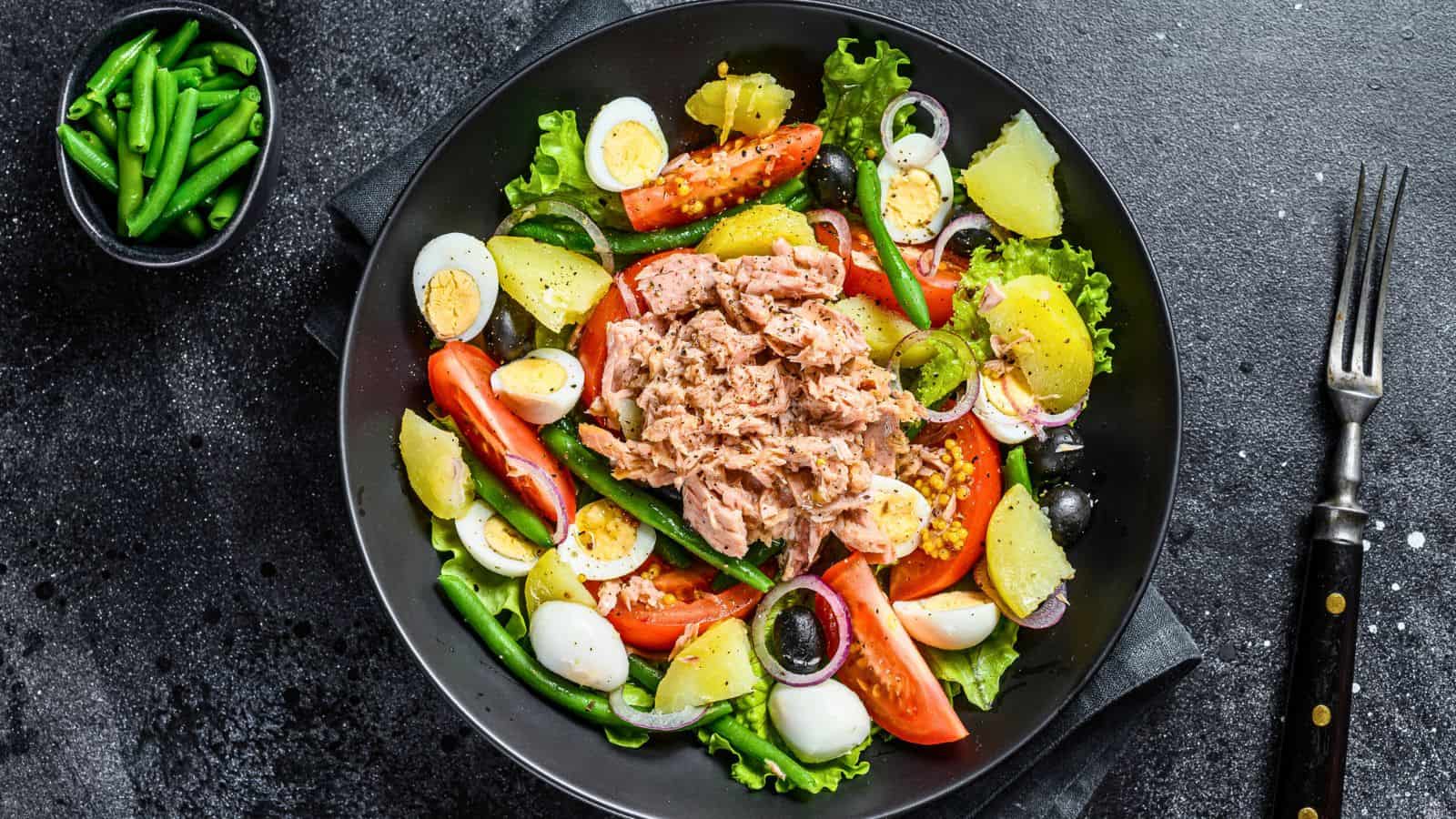 A black plate with a salad including tuna, boiled potatoes, green beans, lettuce, tomato wedges, hard-boiled egg slices, black olives, and onions, next to a small bowl of green beans and a fork on a dark surface.