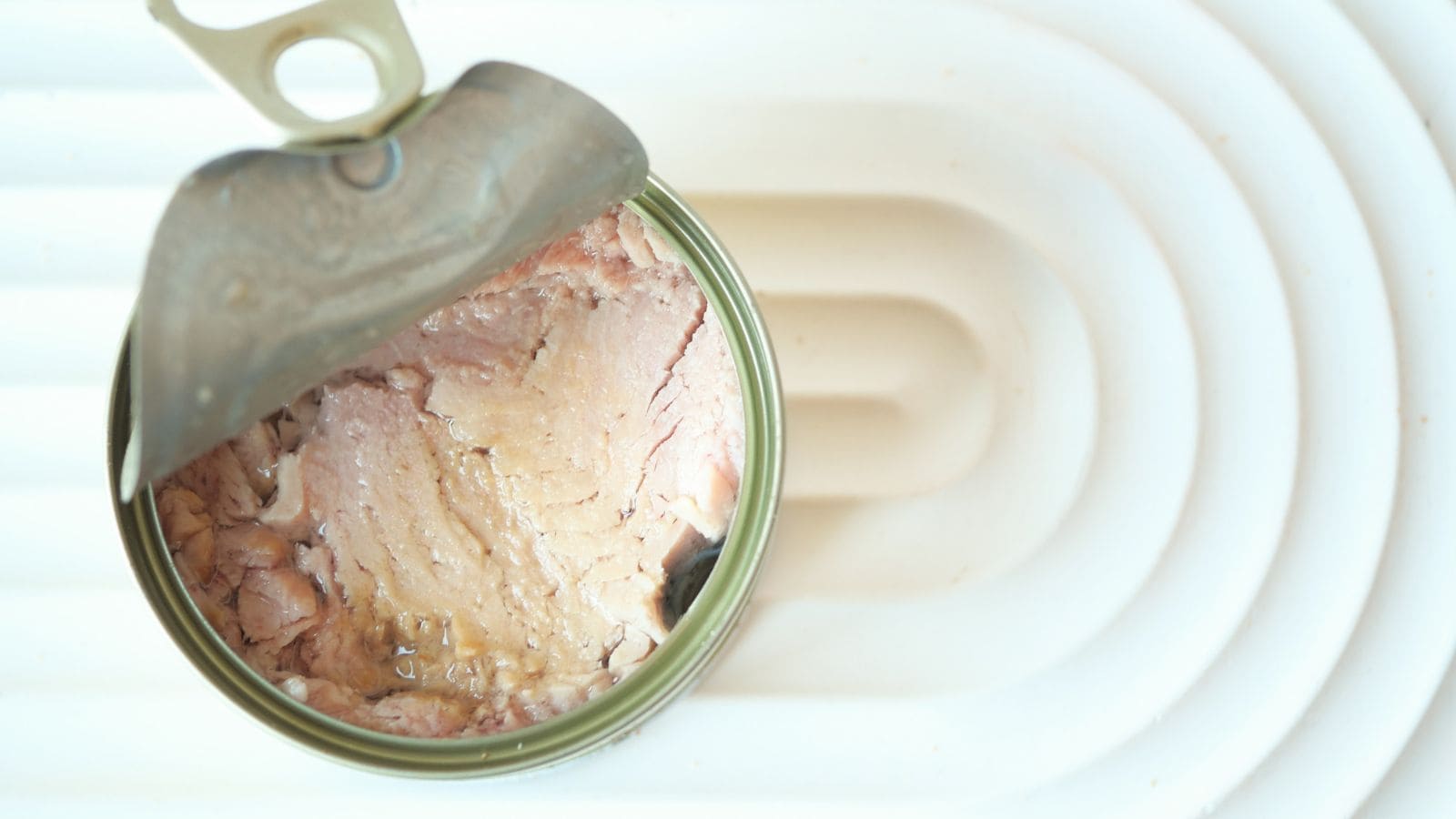 An opened can of tuna sits on a white surface, showing chunks of tuna packed inside with some liquid visible.