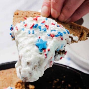 A hand holds a slice of cookie cake topped with white frosting, red, white, and blue sprinkles, and a blue star-shaped decoration. The rest of the cookie cake is visible in a skillet below.
