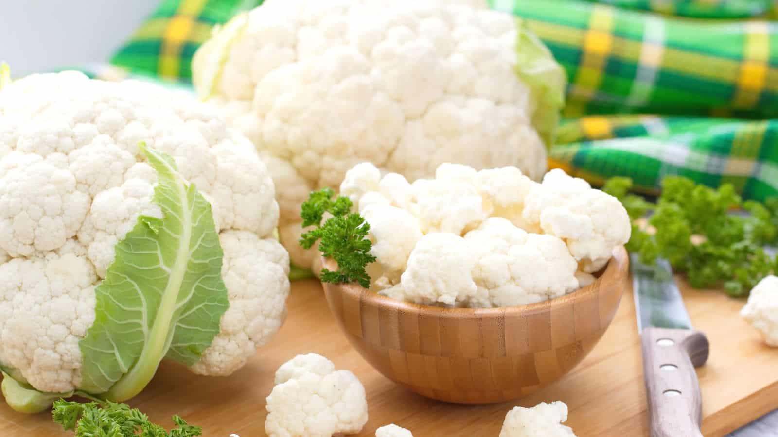 Whole cauliflower heads and a wooden bowl filled with cauliflower florets are arranged on a wooden surface. A knife, sprigs of parsley, and a green checkered cloth are also visible in the background.