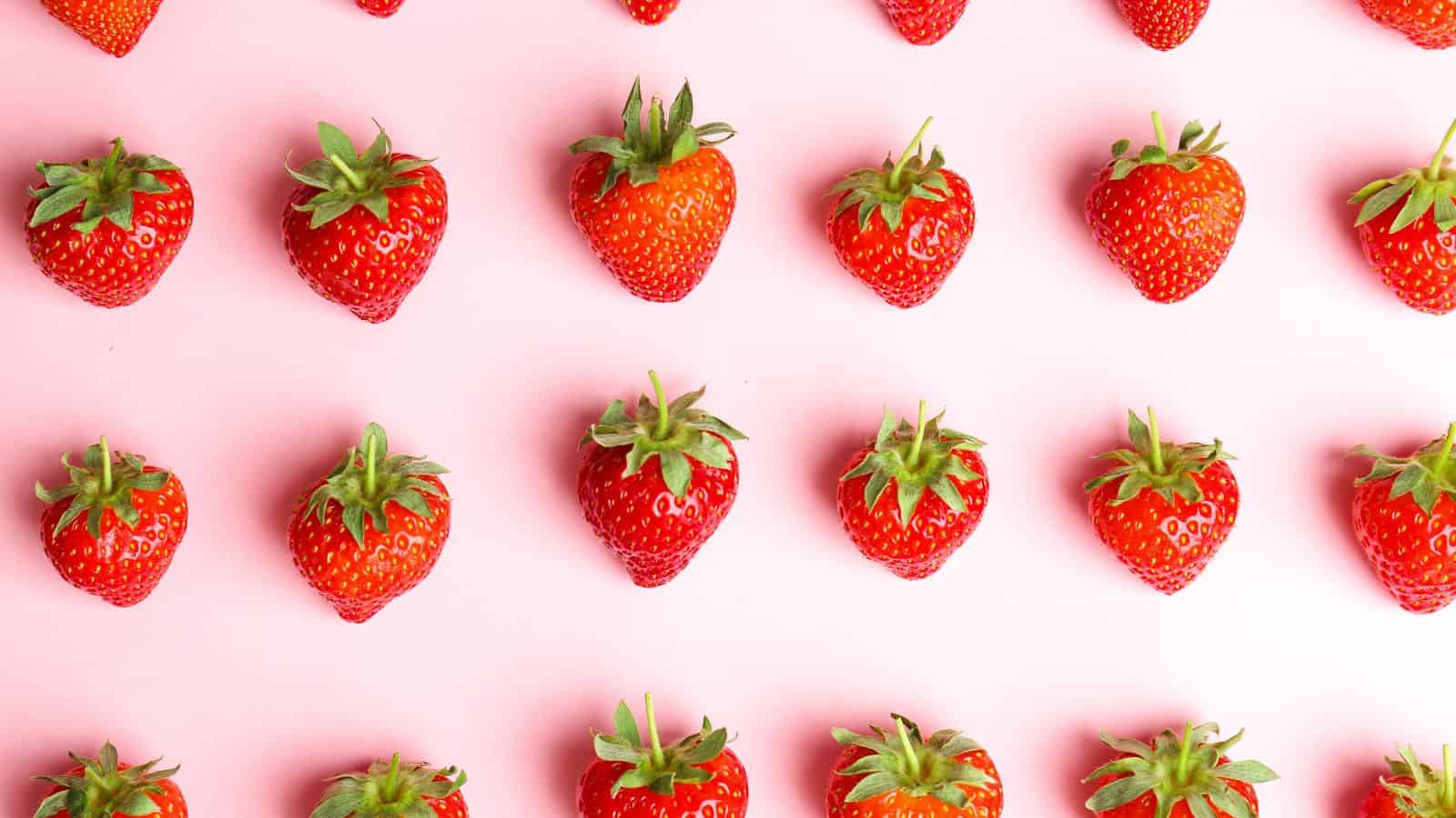 Rows of fresh strawberries with green leaves are evenly spaced on a light pink background. The strawberries are arranged in a regular, repeating pattern.