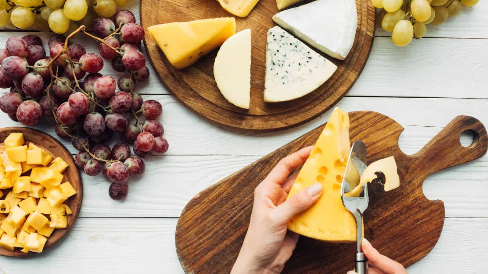 A person slices a wedge of Swiss cheese on a wooden board. Nearby are clusters of grapes, cubes of cheese, and various types of cheese on round wooden platters atop a white wooden table.