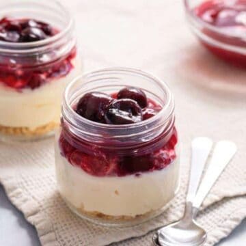 Two small glass jars filled with layered cheesecake dessert topped with cherry compote, placed on a beige cloth. A small glass bowl with extra cherry topping and two metal spoons are nearby.