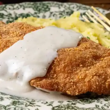 A plate with breaded and fried chicken fried steak topped with white gravy, served next to mashed potatoes. A fork and knife are placed on the patterned plate.