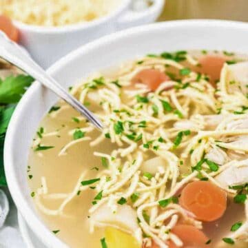 A bowl of chicken noodle soup with sliced carrots, noodles, and chunks of chicken, garnished with chopped parsley. A spoon rests in the bowl, with fresh parsley and a small dish of cheese nearby.