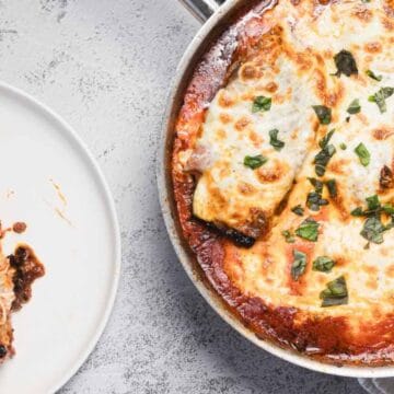A skillet with two pieces of baked eggplant parmesan covered in melted cheese and tomato sauce, topped with fresh basil, sits beside a white plate with a serving of the dish.