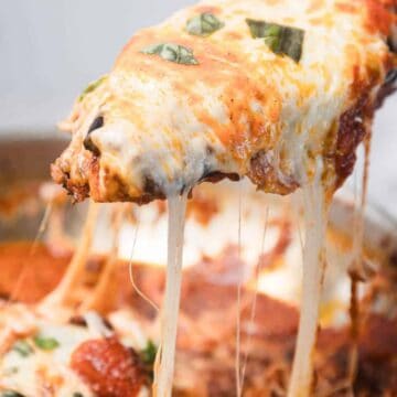 A close-up of a cheesy slice of baked lasagna being lifted from a pan, with melted cheese stretching and visible tomato sauce and basil on top.
