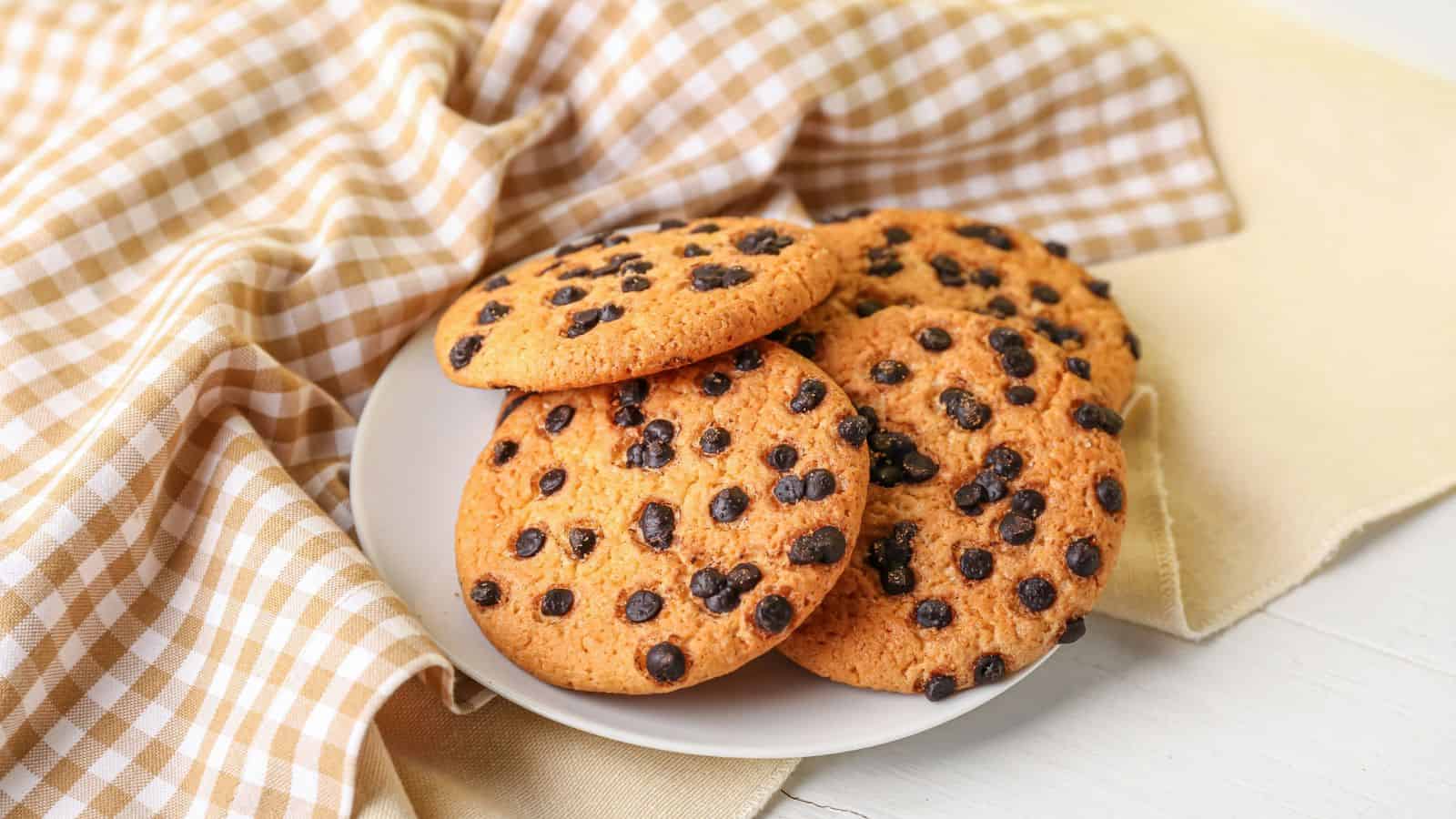 A white plate holds four chocolate chip cookies, placed on a surface next to a beige and white checkered cloth.