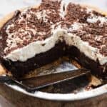 A gluten-free chocolate pie with a thick chocolate filling, topped with whipped cream and chocolate shavings, sits in a foil pie tin with a slice removed. A pie server rests under the pie in the empty space.