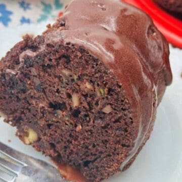 A close-up of a slice of chocolate bundt cake with visible bits of nuts, topped with chocolate glaze, served on a white plate with a metal fork. The background shows a floral-patterned cloth and part of a red platter.