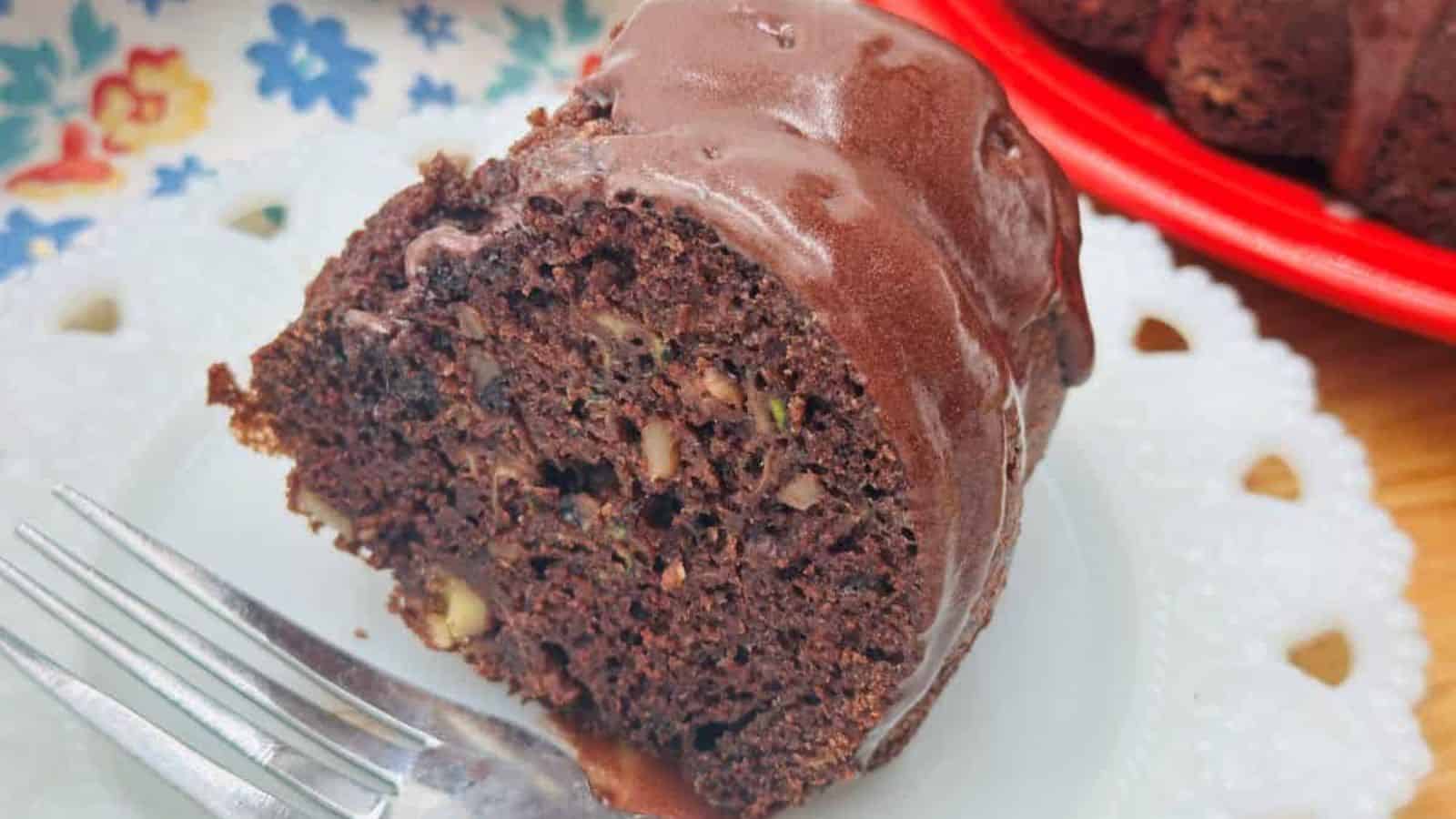 A slice of chocolate bundt cake with walnuts on a white plate, accompanied by a fork. The cake is partially seen on a red plate in the background, with a floral napkin next to it.