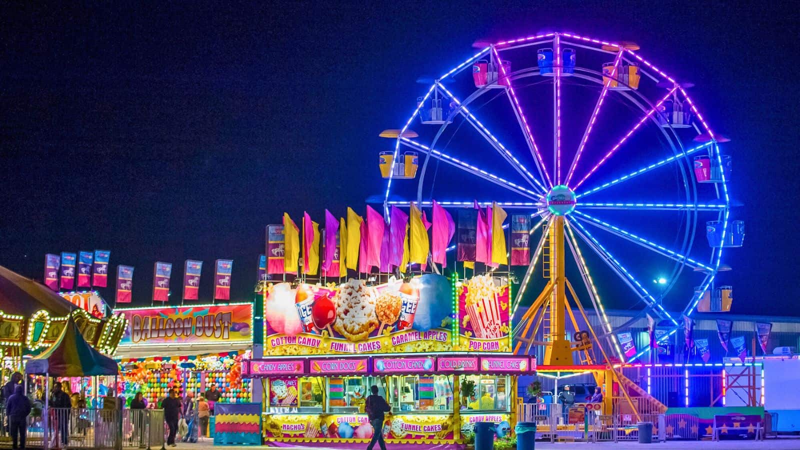 A brightly lit Ferris wheel and colorful carnival booths with food and games are shown at night. Neon lights illuminate the area, and flags decorate the booths. People are visible enjoying the carnival atmosphere.