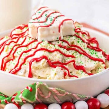 A bowl of holiday dessert featuring a creamy mixture topped with red and green sprinkles. A white chocolate-covered treat rests on top, drizzled with red icing. Candy beads and a festive napkin are in the background. Glass of milk is partially visible.