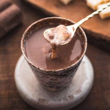 A spoon holding a thick, creamy chocolate mixture hovers over a cup filled with hot chocolate, set on a coaster on a wooden table. Chocolate blocks and a cutting board are visible in the background.