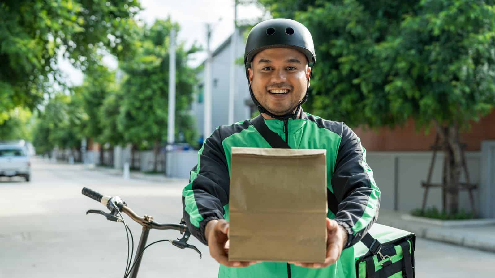 A delivery person wearing a green and black jacket and a helmet stands outdoors next to a bicycle, smiling and holding out a brown paper bag. Trees and houses line the street in the background.