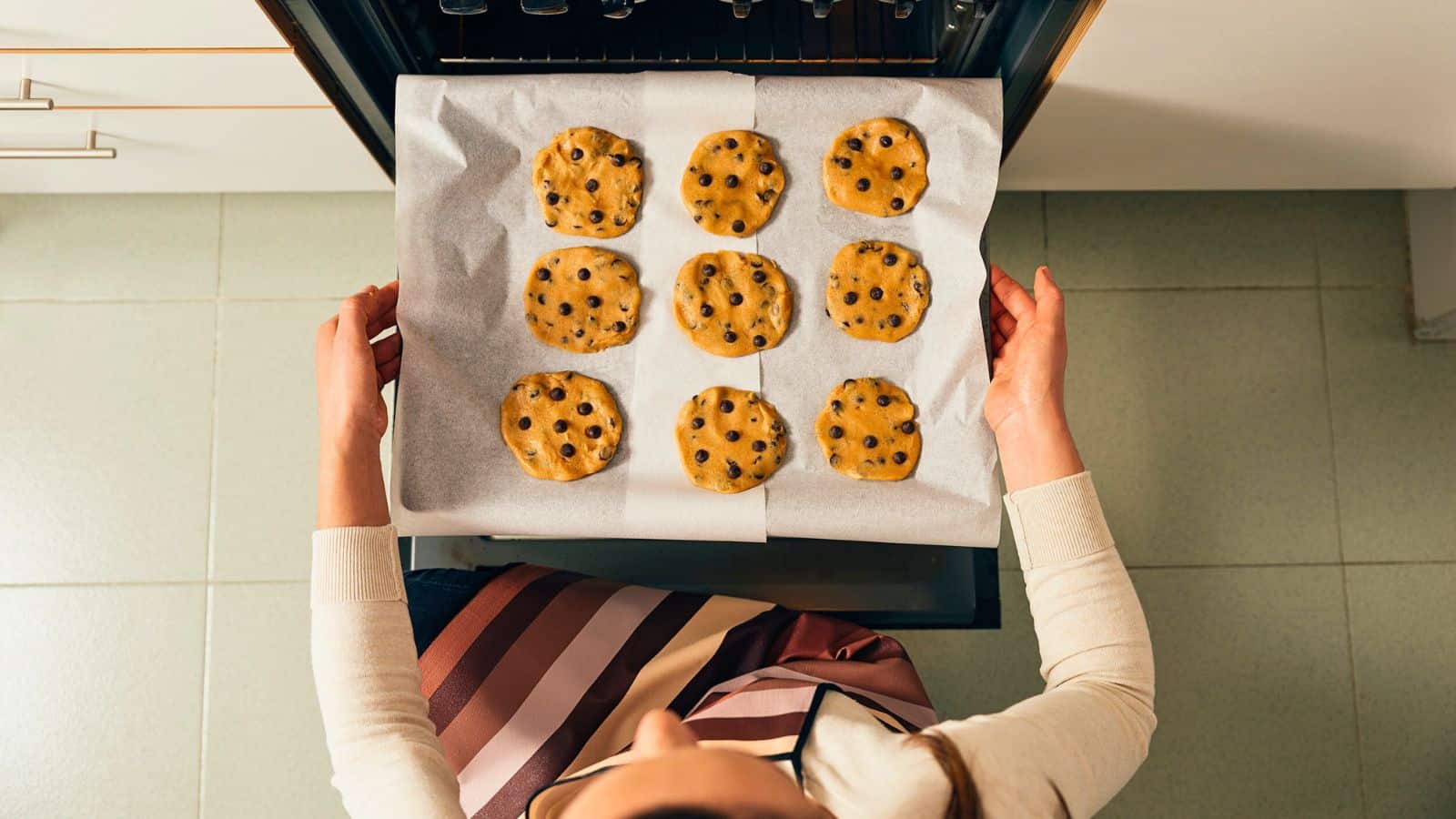 A person places a baking tray lined with parchment paper and nine unbaked chocolate chip cookies into an oven, viewed from above. The person is wearing a striped apron and standing on a light tiled floor.