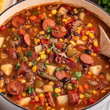 A pot of mixed vegetable and sausage stew with corn, beans, potatoes, and herbs, accompanied by sliced cornbread on the side.