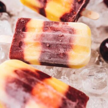 Three striped popsicles in shades of yellow, orange, and purple rest on ice cubes, surrounded by several whole cherries on a light surface.