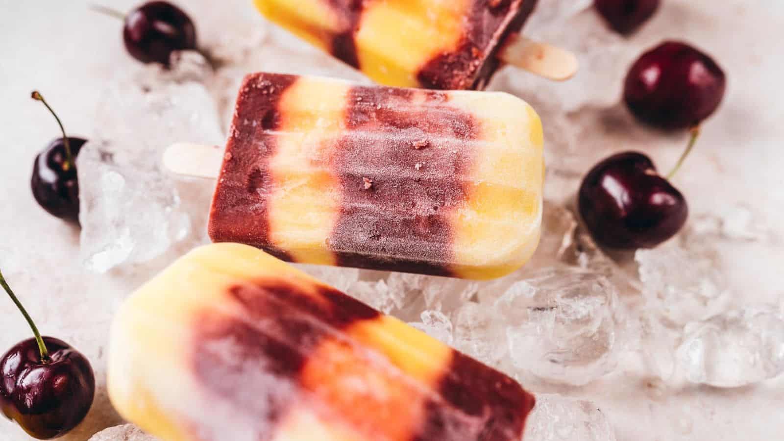 Three striped popsicles in shades of yellow, orange, and purple rest on ice cubes, surrounded by several whole cherries on a light surface.