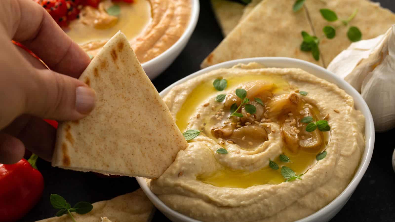 A hand dips a piece of pita bread into a bowl of hummus topped with olive oil, garlic, and herbs. Nearby are another bowl of dip, pita slices, garlic bulbs, and red peppers.