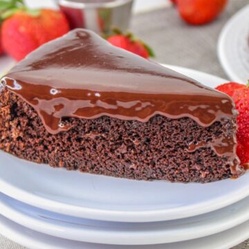 A slice of chocolate cake with glossy chocolate icing sits on stacked white plates, garnished with a fresh strawberry. Whole strawberries and the remaining cake are visible in the background.