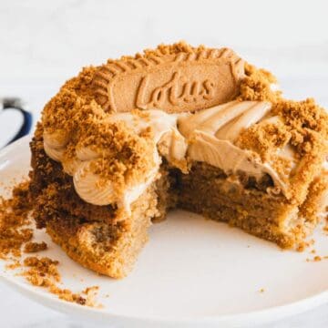 A cake topped with cream, crushed biscuit pieces, and a whole Lotus biscuit sits on a white cake stand. A slice is missing, and a dark blue cup with a silver rim is in the background.