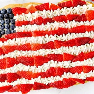 A rectangular sheet cake decorated to resemble the American flag, with blueberries in the top left corner and alternating rows of sliced strawberries and white frosting covering the rest of the cake.