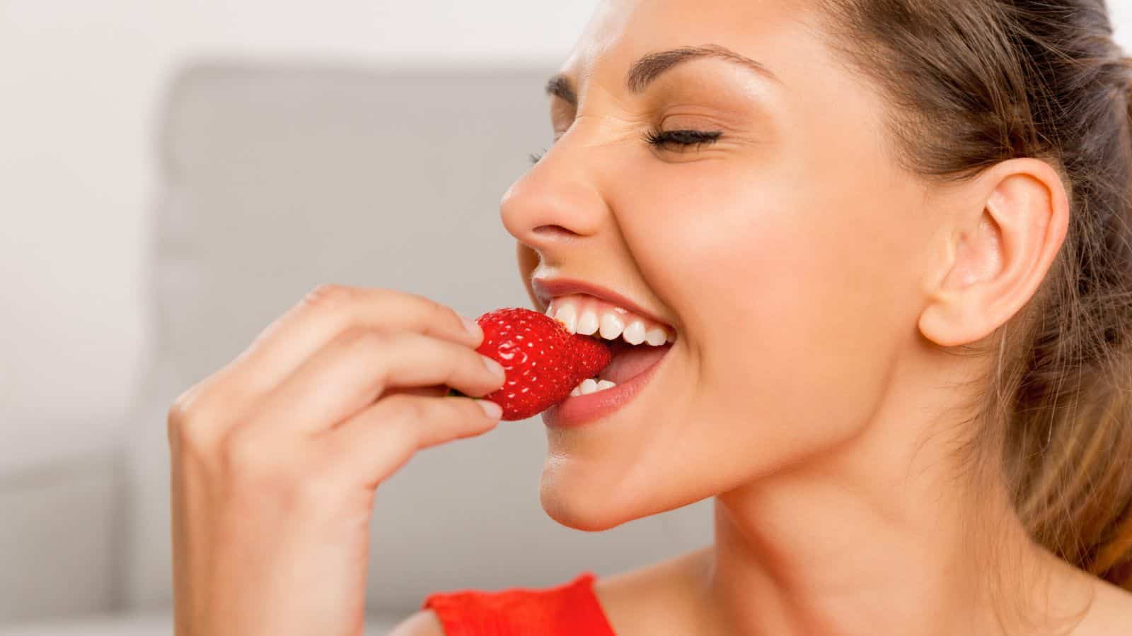 A woman in a red top holds a strawberry close to her mouth and smiles as she prepares to take a bite. The background is out of focus, showing a neutral-colored setting.