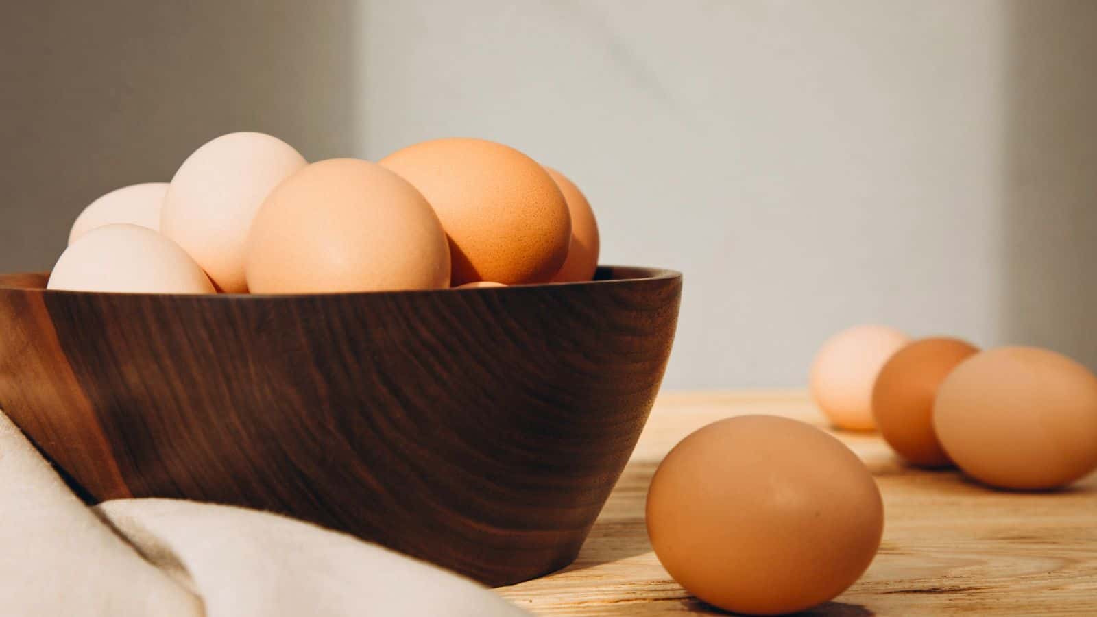 A wooden bowl filled with brown and white eggs sits on a light wooden surface, with a few eggs scattered nearby and a white cloth partially visible in the foreground.