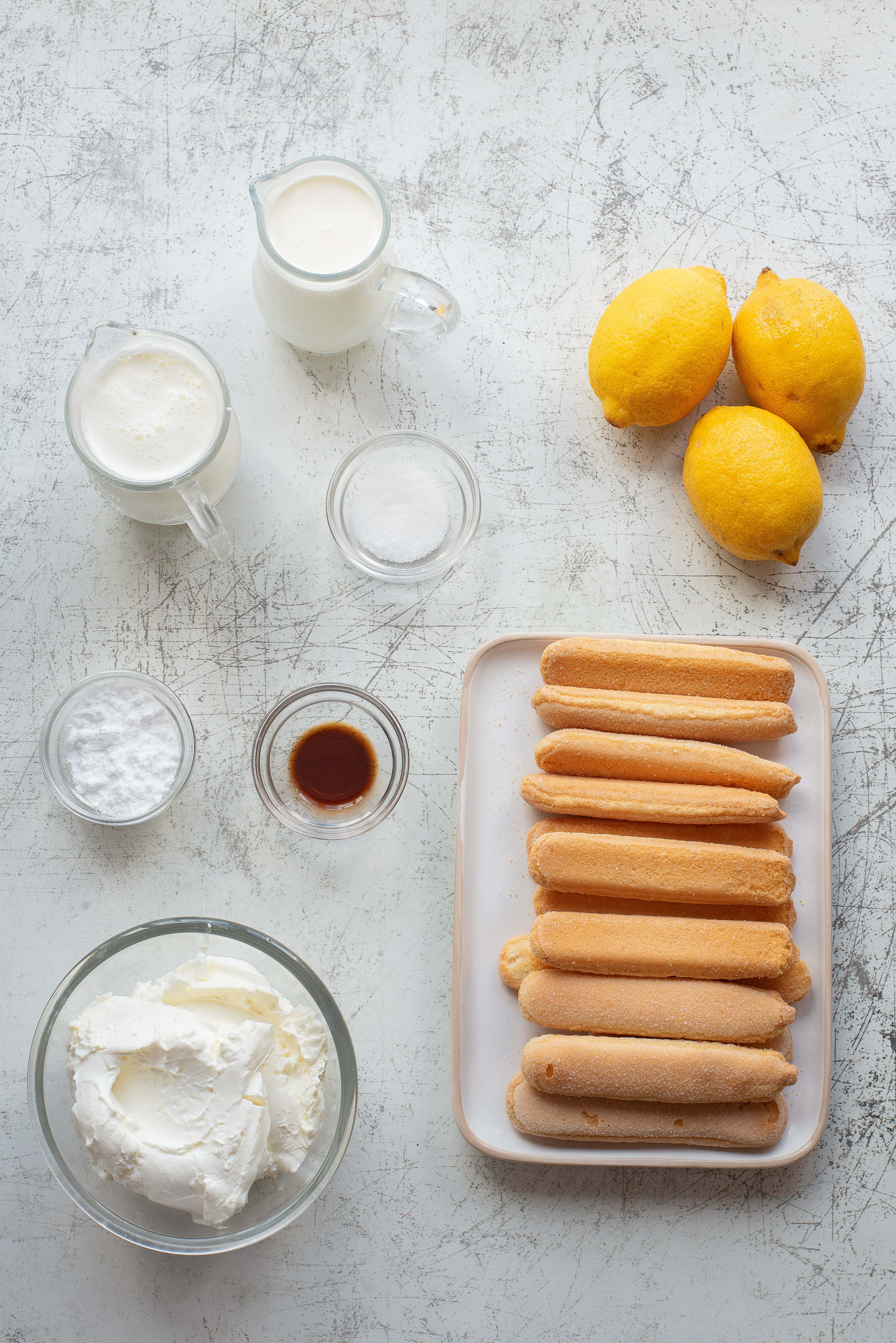 A flat lay of baking ingredients including three lemons, two glass pitchers of milk and cream, a small bowl of powdered sugar, a small bowl of granulated sugar, vanilla extract, mascarpone in a bowl, and ladyfinger cookies on a plate.
