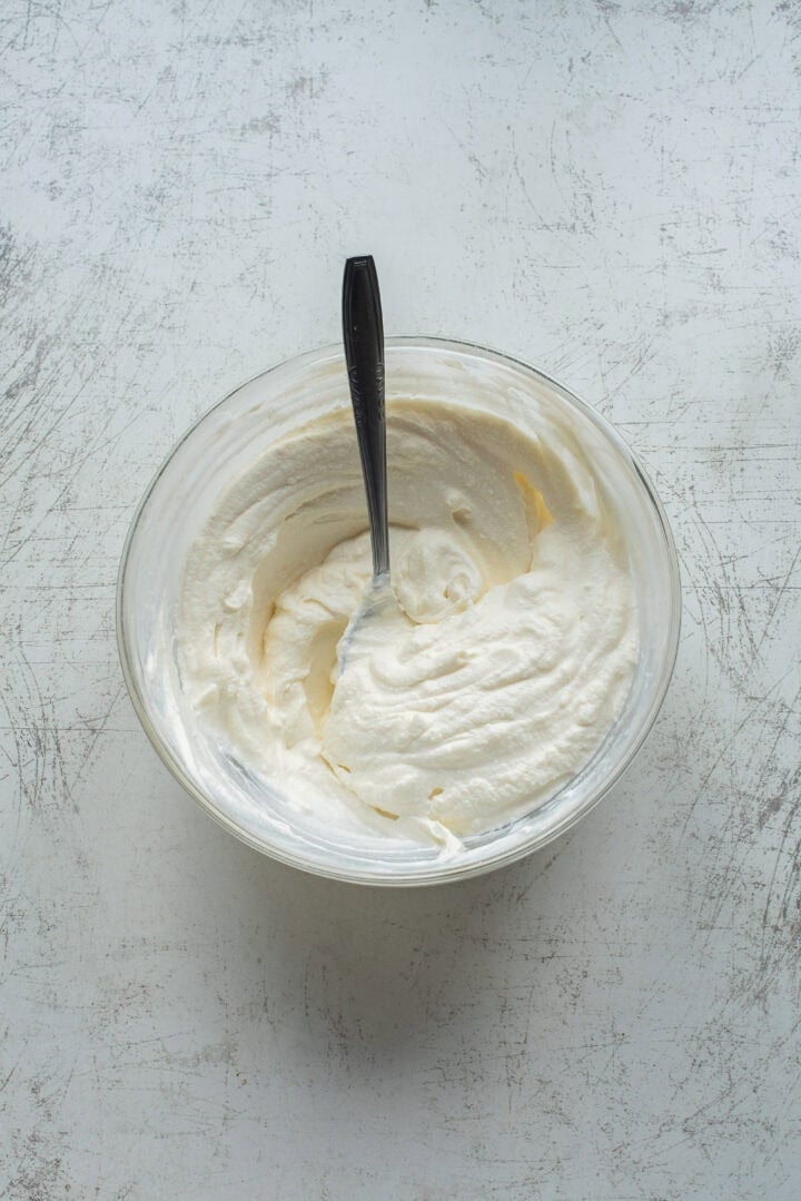 A glass bowl filled with whipped cream, with a metal spoon resting inside, sits on a light-colored textured surface.
