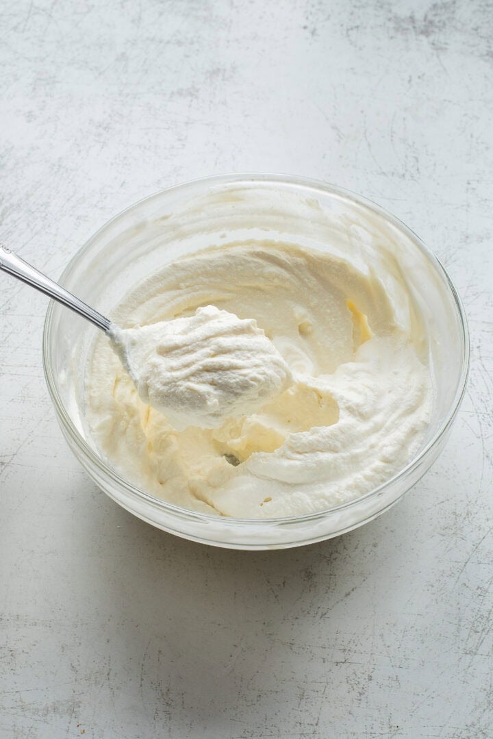 A glass bowl filled with whipped cream sits on a light, textured surface. A metal spoon rests inside the bowl, holding a dollop of the whipped cream above the rest.