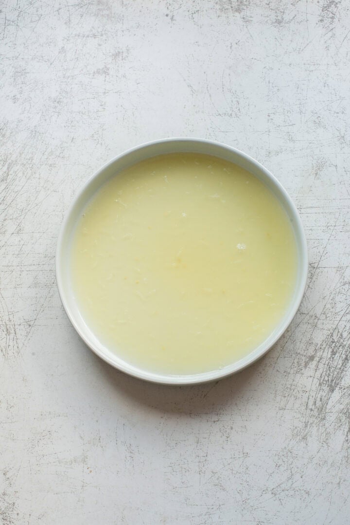 A round white bowl filled with a pale yellow liquid sits on a textured, light-colored surface.