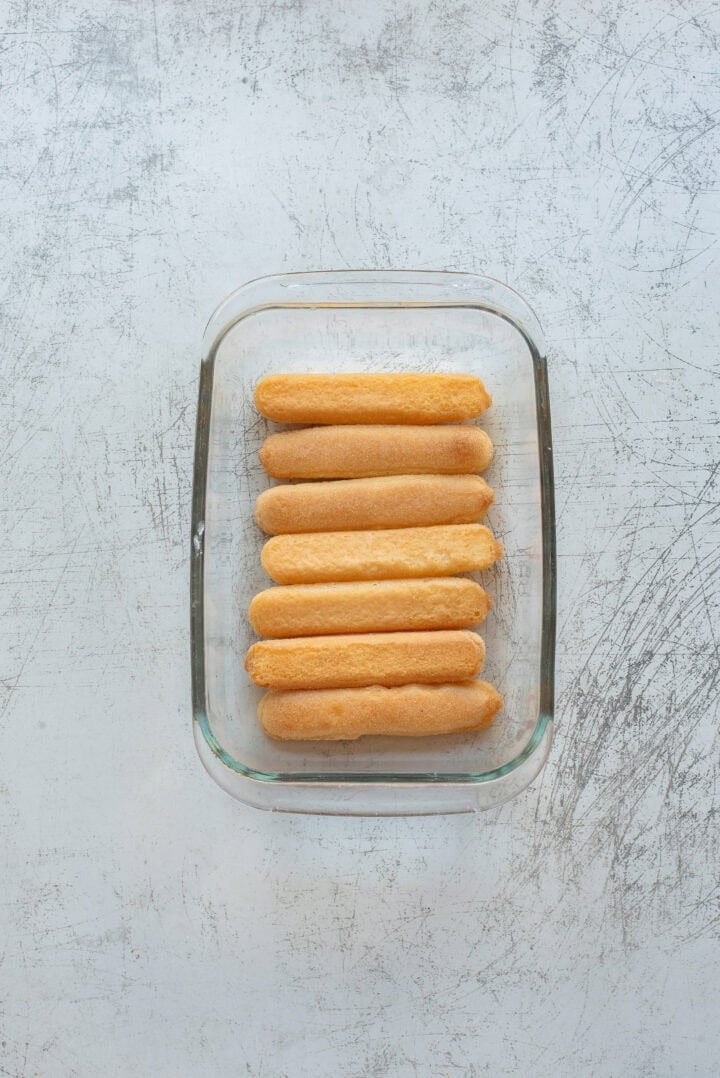 A glass rectangular dish containing seven evenly aligned ladyfinger cookies on a light, textured surface.