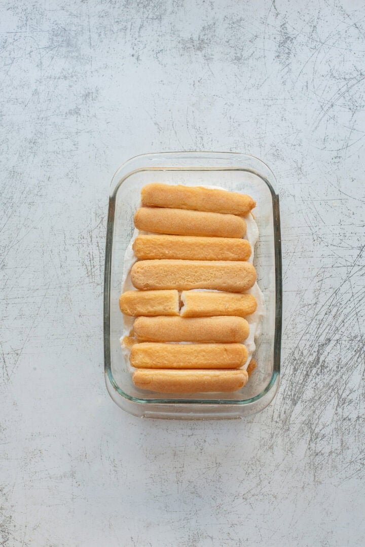 A glass dish with a layer of cream topped with nine ladyfinger biscuits, one of which is broken in half, on a textured light gray surface.