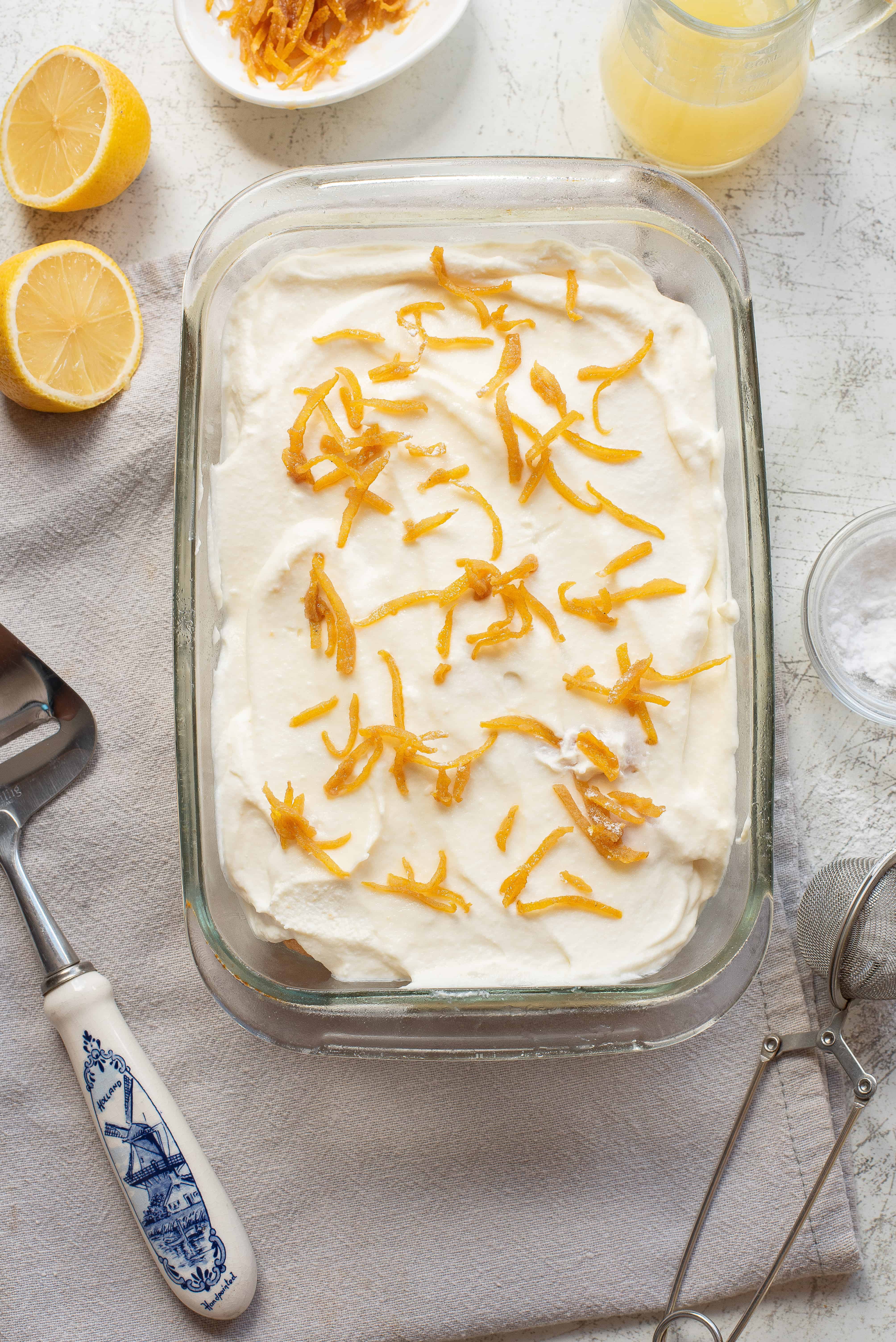 A glass dish filled with a creamy dessert topped with candied lemon peel, surrounded by halved lemons, a dish of more candied peel, a pitcher of liquid, a pie server, and a sifter on a light surface.