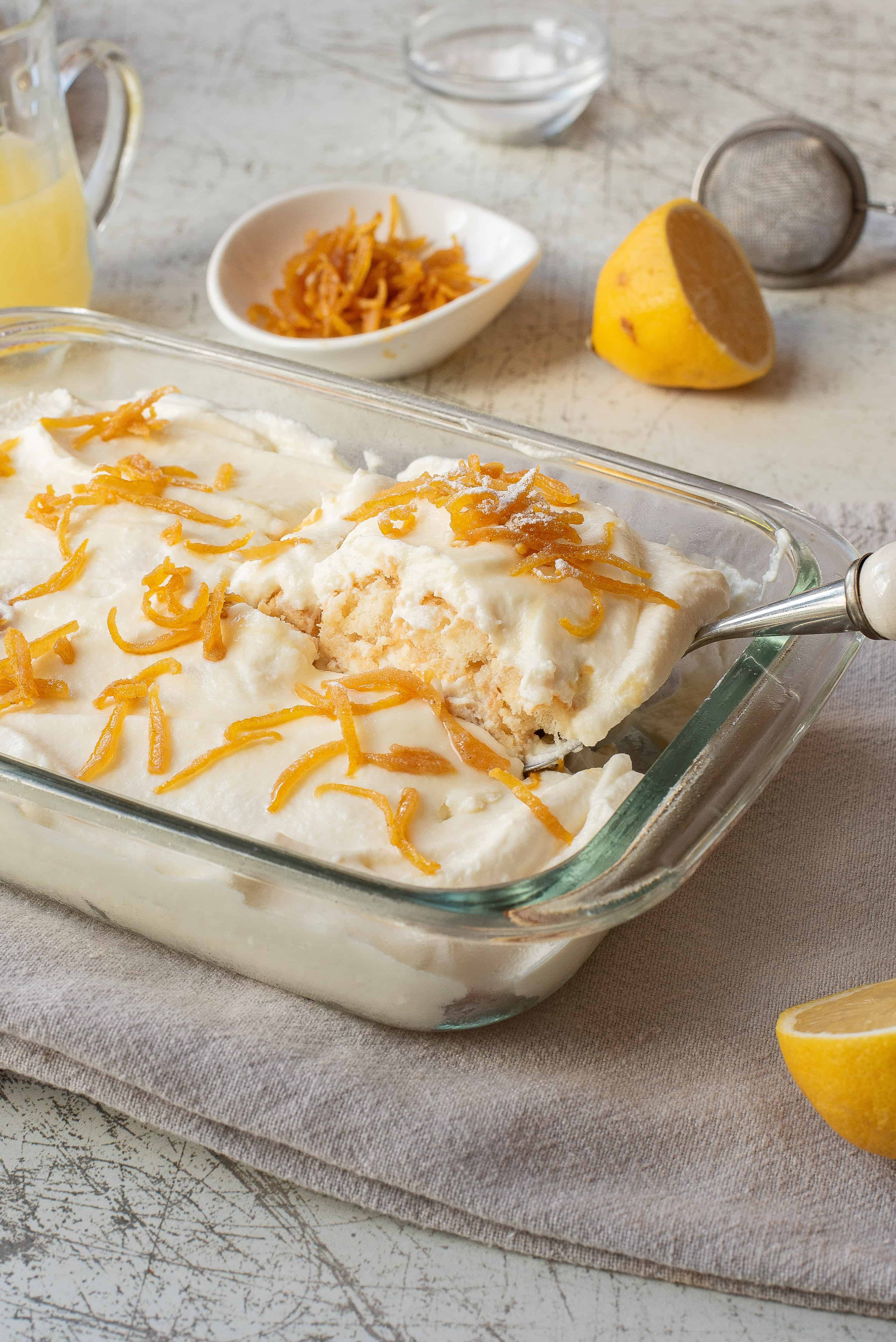 A glass baking dish filled with a creamy dessert topped with thin strips of candied lemon peel. A serving has been lifted out with a spatula. Sliced lemons and a small dish of candied peel are in the background.
